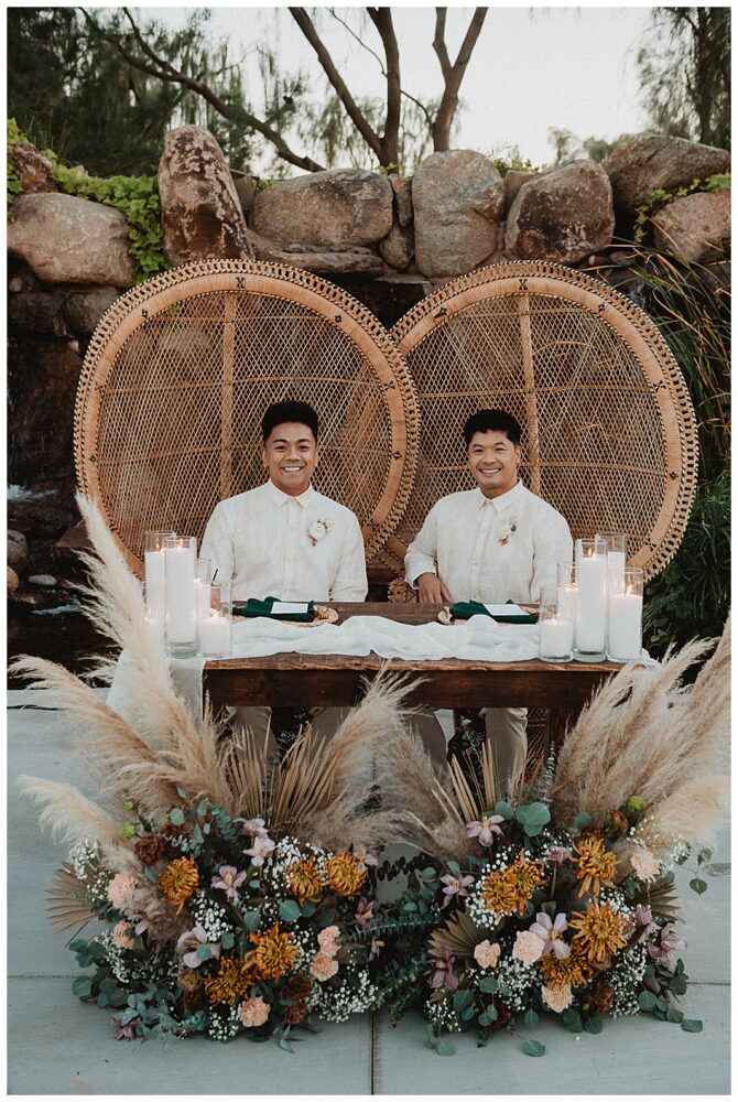 Two grooms seated at decorated wedding table.
