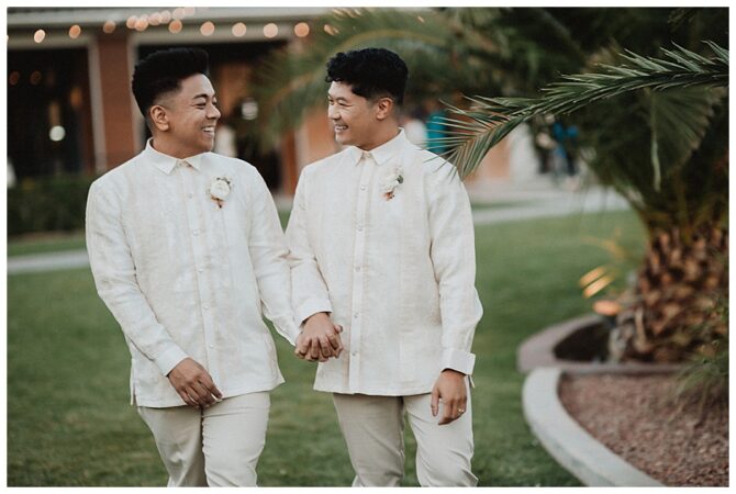 Two grooms holding hands, smiling outdoors.
