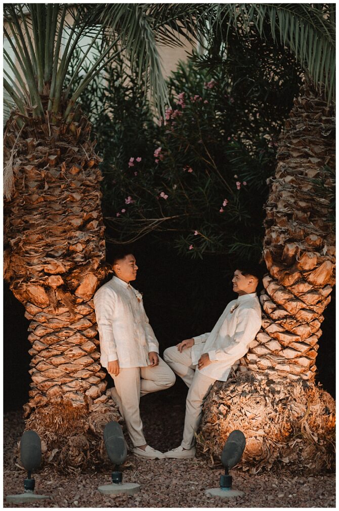 Two people pose between palm trees at night.