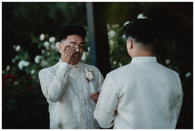 Groom wipes tears during wedding ceremony outdoors.