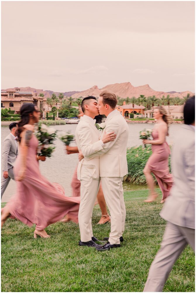 Grooms kissing by lake with bridesmaids passing by.